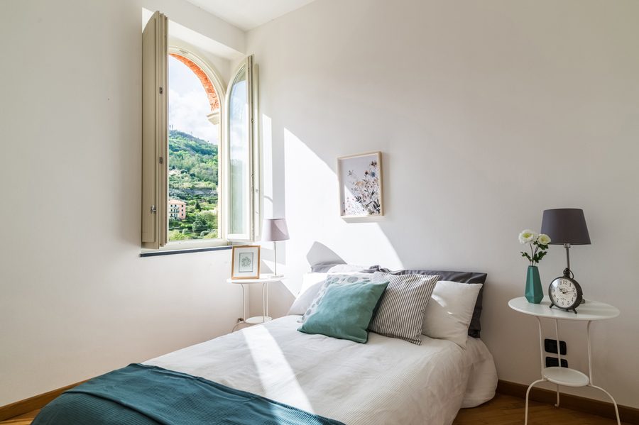 Camera curata dall'home stager: letto con accenti teal e finestra ad arco sulle colline liguri